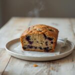 A loaf of freshly baked vegan blueberry banana bread on a wooden table, sliced to reveal moist interior with blueberries.