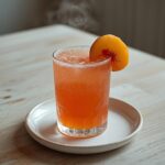 A tall glass of Peach Bellini Mocktail Recipe with a peach slice garnish, showing a bubbly, orange-pink drink.