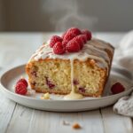 A whole white chocolate raspberry loaf cake on a plate with raspberries and white glaze, ready to serve.